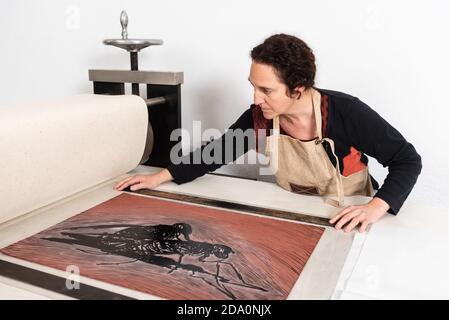 Focused middle aged female master putting sheet of linoleum with inked ornament on printing press while working in linocut technique in work studio Stock Photo