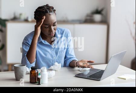 Sick black female office employee sitting at workplace taking lots of pills Stock Photo