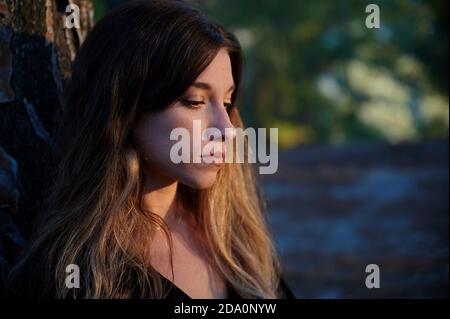 Side view of silent sad young long haired female leaning back on tree trunk while standing against blurred forest Stock Photo