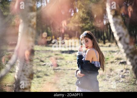 Side view of serene female in denim jacket standing in park on sunny day in summer and looking at camera Stock Photo