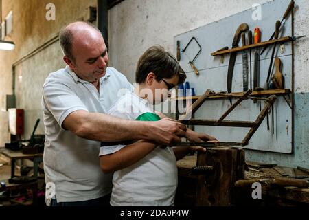 Side view of father teaching son using metal rasp for grinding wood in grungy dirty woodwork workshop Stock Photo