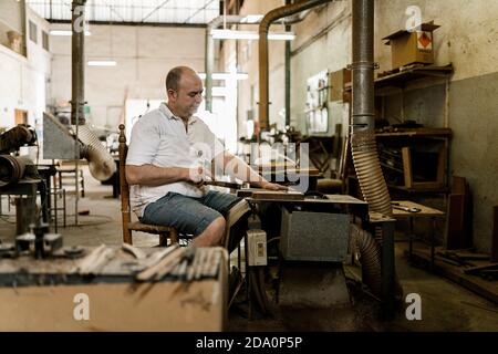 Determined male woodworker sitting at old workbench and working with lumber details in grungy workshop Stock Photo