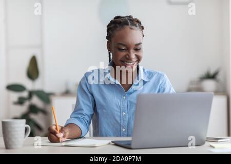 Online Education. Young Black Female Student Using Laptop At Home, Taking Notes Stock Photo