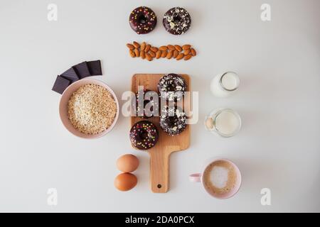 Top view composition with sweet donuts with chocolate topping and sprinkles placed on wooden board on white table with ingredients for recipe Stock Photo