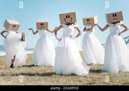 Full body of women wearing bridal dresses and boxes with happy and sad smileys on head standing in field Stock Photo