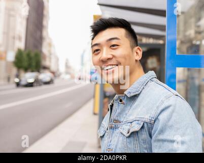 Side view of smiling handsome Asian male in stylish denim clothes standing on street and looking away Stock Photo