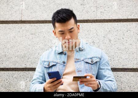 Young Asian male in denim jacket doing online payment with mobile phone and credit card while standing against stone wall on urban street Stock Photo