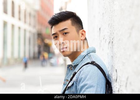 Side view of serious young Asian male student in denim jacket with piercing in ear standing near wall of urban building and looking at camera Stock Photo