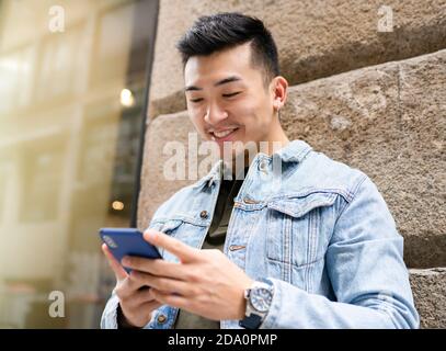 Content young Asian male in casual outfit browsing on mobile phone while standing near stone wall of urban building Stock Photo