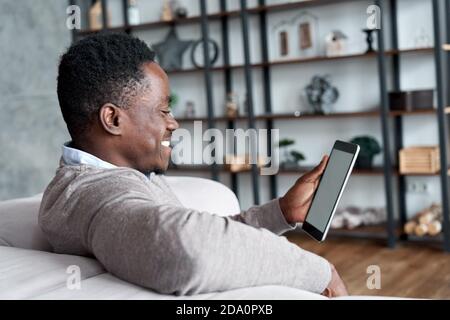 Happy african man using digital tablet apps sitting on couch at home. Stock Photo