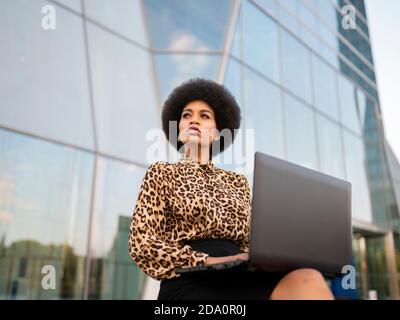From below busy African American female with afro sitting on bench with computer typing text while working remotely near glass building Stock Photo