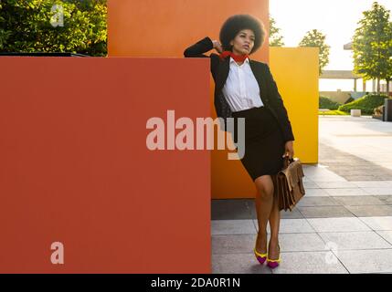 Full body of elegant African American businessWoman in classy suit leaning on red cube and thoughtfully looking away Stock Photo