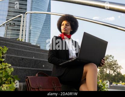 From below serious African American entrepreneur with afro hair working on laptop sitting on stairs looking away Stock Photo