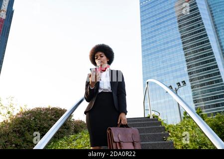 Low angle serious African American female entrepreneur in formal wear with briefcase using mobile phone on street Stock Photo