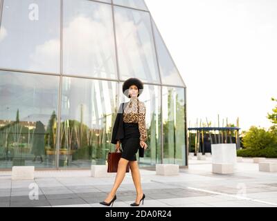 Full body of unemotional thoughtful stylish African American female employee with afro hair and briefcase walking near glass walled building Stock Photo