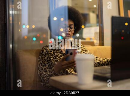 Through glass of serious African American female scrolling cellphone while working remotely at table with laptop in cafe Stock Photo