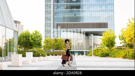 Full body of unemotional thoughtful stylish African American female employee with afro hair and briefcase walking near glass walled building Stock Photo