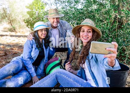 Cheerful people sitting together and taking selfie on smartphone while resting after harvest in garden Stock Photo