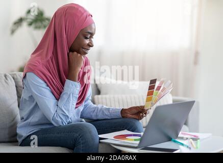 Smiling Black Muslim Lady Holding Color Swatch And Using Laptop At Home Stock Photo
