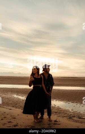 Full body of young barefoot man and Woman in casual clothing holding hands and looking in opposite directions while standing on wet sand covered with Stock Photo