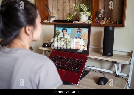 Young Asian female sitting on desk chatting via video call on laptop with group of friends while spending time at home during coronavirus pandemic Stock Photo