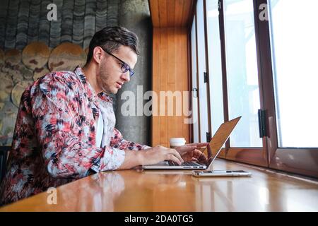 Young focused male freelancer with eyewear and disposable glass of hot drink sitting at windowsill with netbook Stock Photo