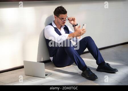 From above of young masculine office worker in formal suit and eyewear sitting on floor near wall with cellphone and netbook Stock Photo