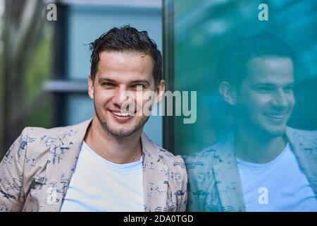 Young glad office worker in trendy clothes with cellphone looking at camera while leaning on wall in city Stock Photo