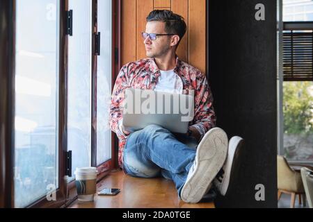 Young trendy remote worker in eyeglasses with netbook sitting with crossed legs on windowsill near disposable glass of coffee while looking out window Stock Photo