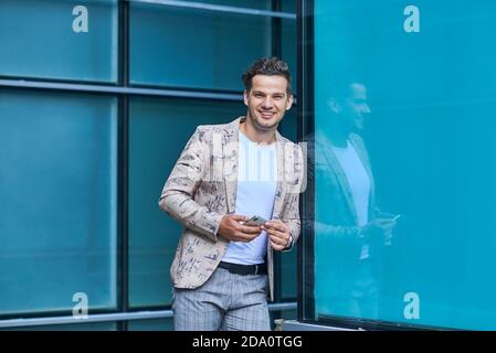 Young glad office worker in trendy clothes with cellphone looking at camera while leaning on wall in city Stock Photo