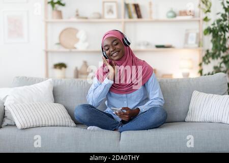 Black Muslim Woman Wearing Wireless Headphones, Listening Music On Smartphone At Home Stock Photo