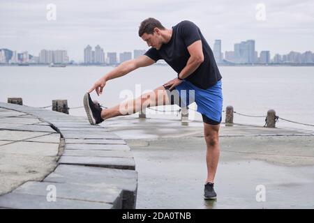Concentrated male runner in sportswear standing on embankment and warming up while stretching legs and doing forward bend during workout Stock Photo