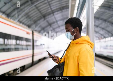 Ethnic male traveler with backpack and in mask standing on platform with train and using cellphone while waiting for departure during COVID 19 epidemi Stock Photo