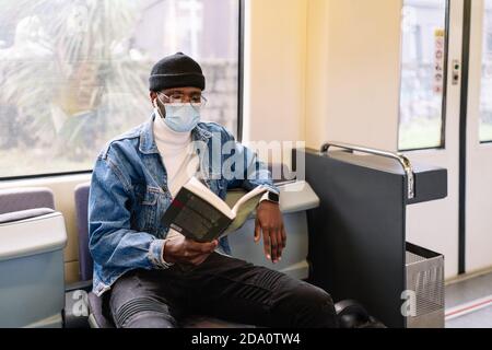 Serious African American male in medical mask sitting on passenger seat in modern train and reading book during coronavirus pandemic Stock Photo