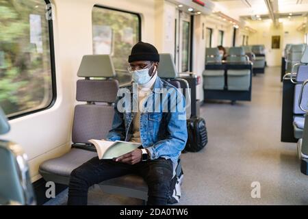Serious African American male in medical mask sitting on passenger seat in modern train and reading book during coronavirus pandemic Stock Photo