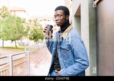 Handsome African American male in denim clothes sitting on stool on street and drinking delicious coffee to go while looking at camera Stock Photo
