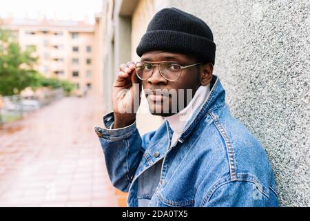 Side view of African American male in trendy denim jacket putting on eyeglasses while standing on street near urban building and looking at camera Stock Photo