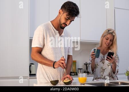 man with fresh avocado and orange juice in glass standing in kitchen with Woman using smartphone and drinking coffee at breakfast Stock Photo