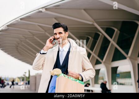 Positive man with black hair in formal clothes standing with shopping bags and talking on phone against modern building Stock Photo