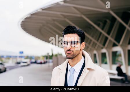 Thoughtful man with black hair and beard in glasses and formal clothes standing on street against modern building at daytime Stock Photo