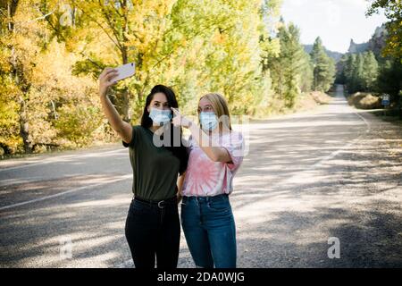 Cheerful young female friends in casual outfits and protective masks taking selfie on smartphone while standing on asphalt road passing through forest Stock Photo