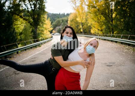 Cheerful young female friends in casual wear and protective masks hugging and having fun while standing on asphalt country road leading through forest Stock Photo