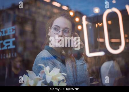 Through window of dreamy female in trendy outfit resting in cozy cafe in evening and looking away while enjoying weekend Stock Photo