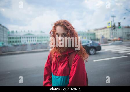 Serious millennial redhead female with long curly hair wearing red jacket looking at camera while standing alone against road and historic buildings i Stock Photo
