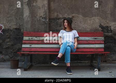 Full body of stylish young Woman in informal outfit and hat resting on old bench against aged stone building in city Stock Photo