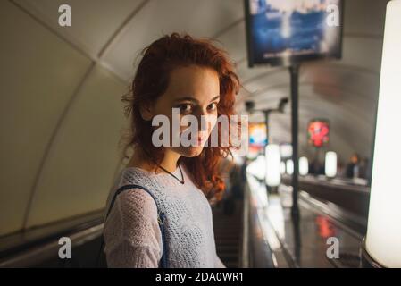 High angle side view of young female passenger in casual outfit with red curly hair looking at camera with smile while going down subway escalator in Stock Photo