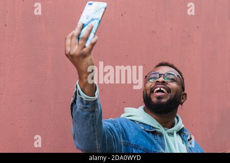 Low angle cheerful adult black hipster guy in casual outfit taking self portrait on mobile phone against red background Stock Photo