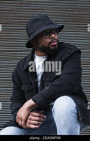 African American bearded guy with missing tooth in eyeglasses wearing trendy casual outfit and hat looking away while sitting against shuttered wall Stock Photo