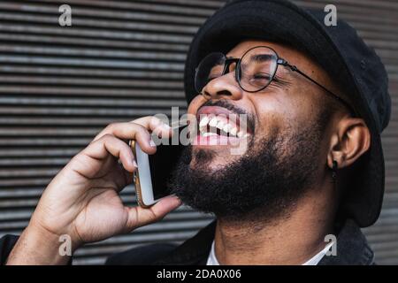 Cheerful adult African American hipster male in hat and eyeglasses talking on mobile phone while standing near metal wall on street Stock Photo