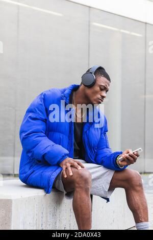 Calm ethnic male wearing stylish hipster outfit sitting on stone border on street in headphones and using mobile phone while looking away Stock Photo